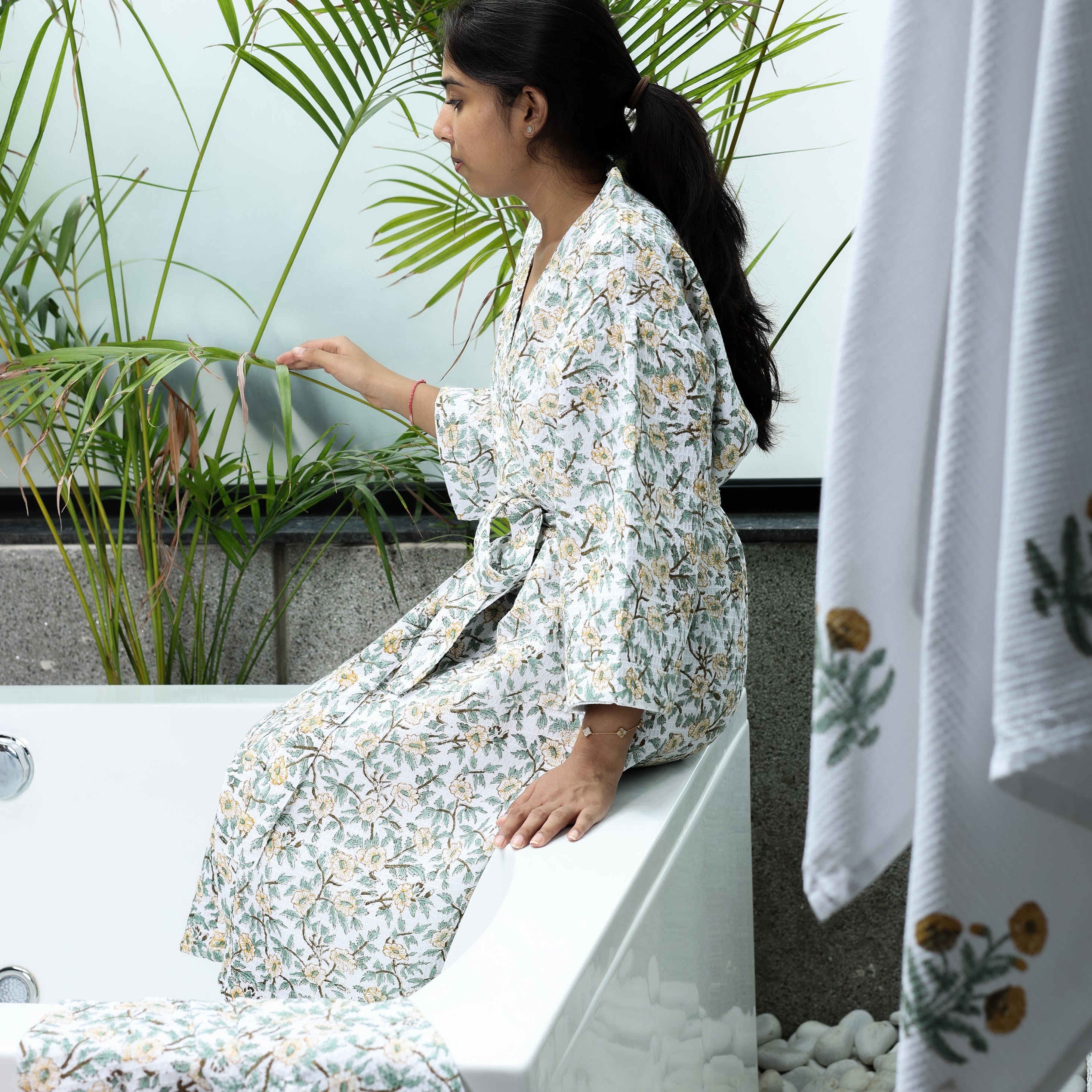 Woman in a floral cotton bath robe sitting on a bench with plants and a towel in the foreground. Bath robes for couples.