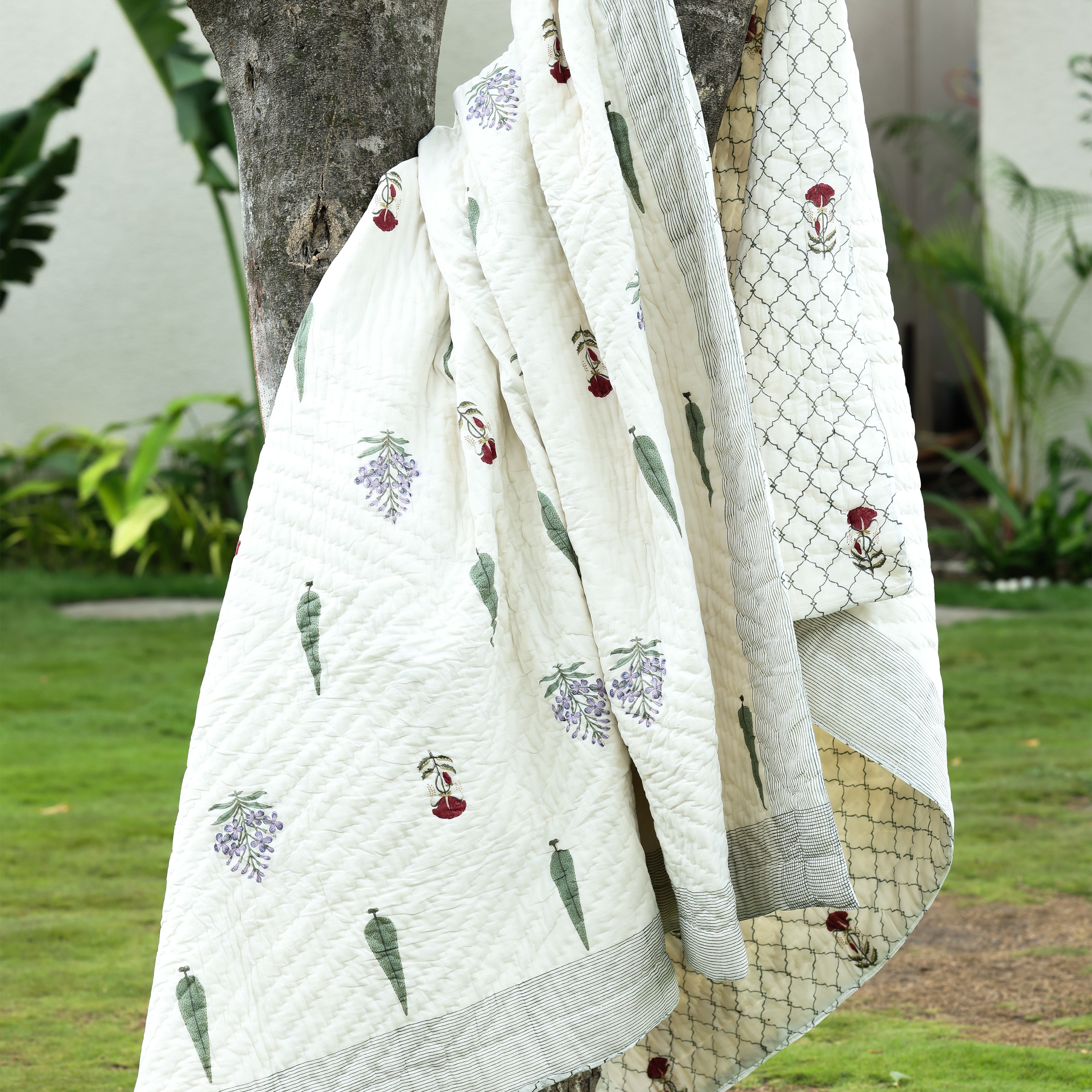 White AC quilt with floral patterns draped over a tree in a garden setting, featuring the brand 'mish mash'. Buy jaipur razai.