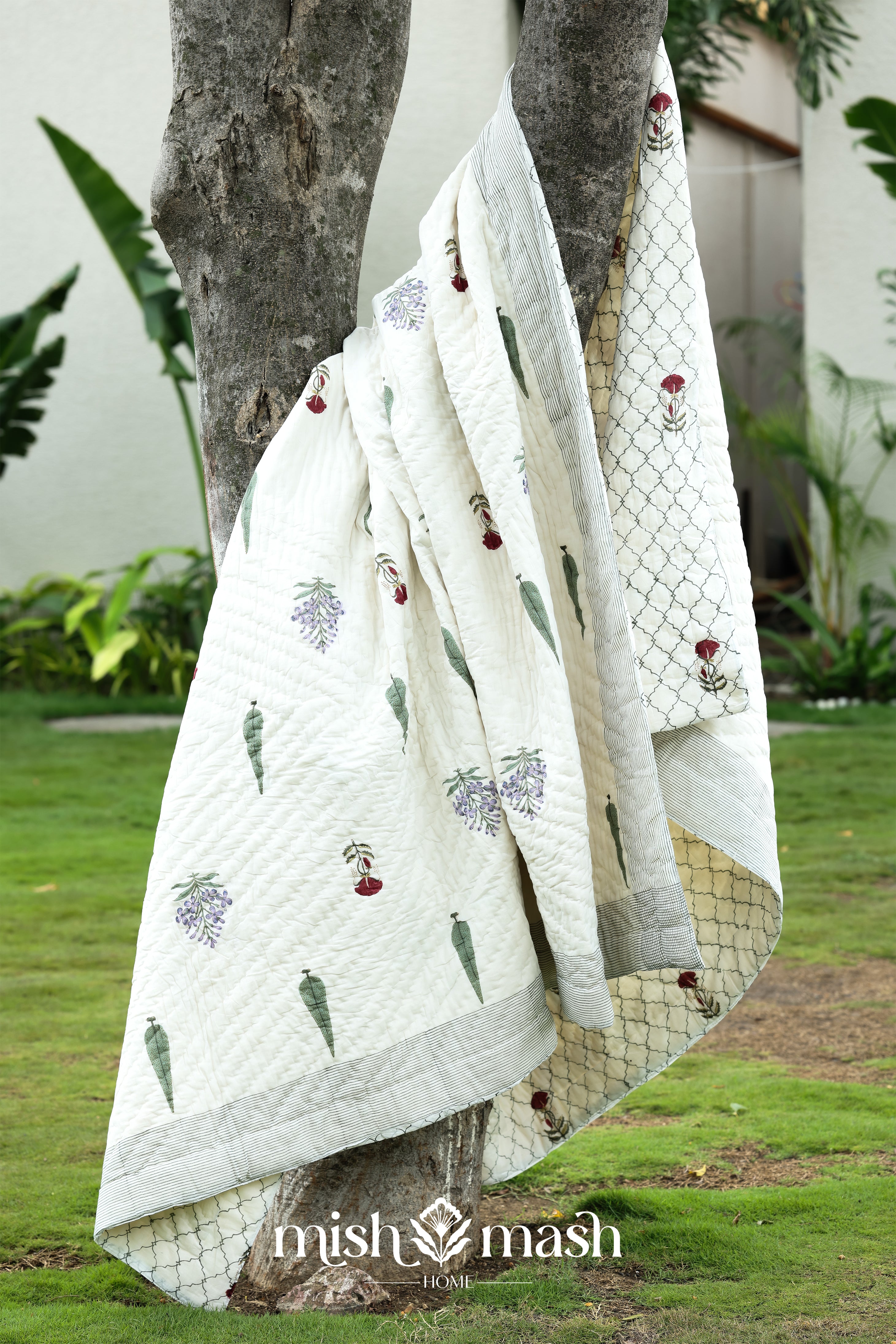 White AC quilt with floral patterns draped over a tree in a garden setting, featuring the brand 'mish mash'. Buy jaipur razai.