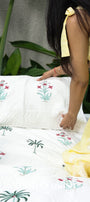Person in a yellow outfit arranging floral  pillows on a cotton bed cover in white. Buy housewarming gifts online. 