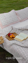 Outdoor setting with a red and grey cotton bed spread, pillows, and a book on a grassy background, in a picnic setup