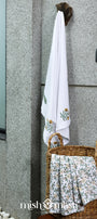 White cotton towel with floral block print hanging on a wall next to a woven basket with 2 folded hand towels.