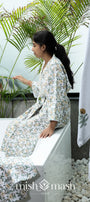Woman in a floral cotton bath robe sitting on a tub with plants and a bath towel and 2 hand towels in the foreground.
