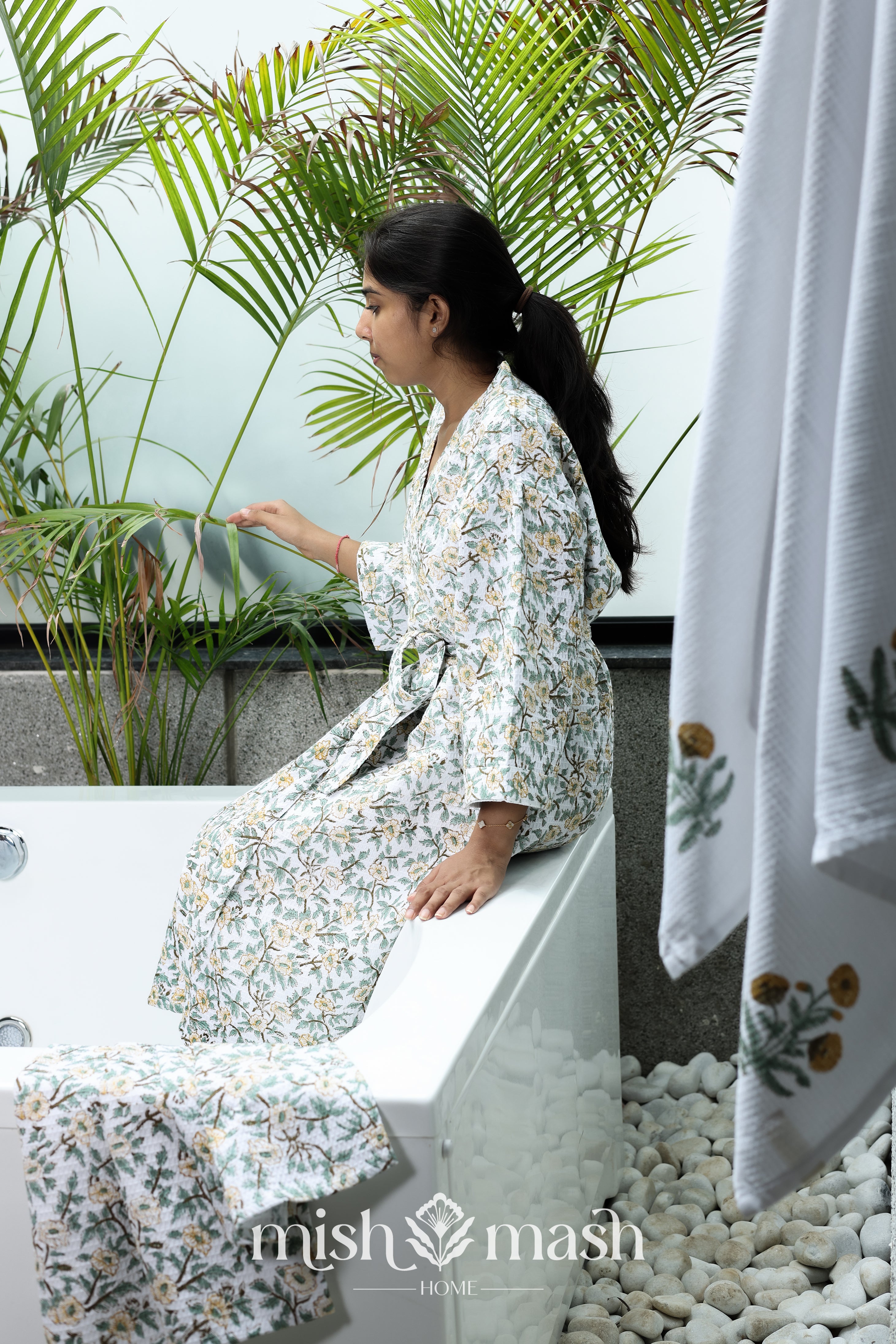 Woman in a floral cotton bath robe sitting on a tub with plants and a bath towel and 2 hand towels in the foreground.