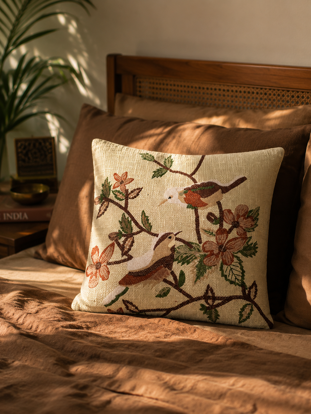A square decorative cushion cover with a detailed embroidered design of birds and flowers on a beige jute background, styled on a bed with brown bedding and wooden headboard in warm natural light.