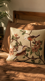 A square decorative cushion cover with a detailed embroidered design of birds and flowers on a beige jute background, styled on a bed with brown bedding and wooden headboard in warm natural light.