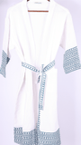 A white cotton bathrobe with blue geometric patterned trim on the cuffs, hem, and tie belt, hanging on a wooden hanger against a light background.