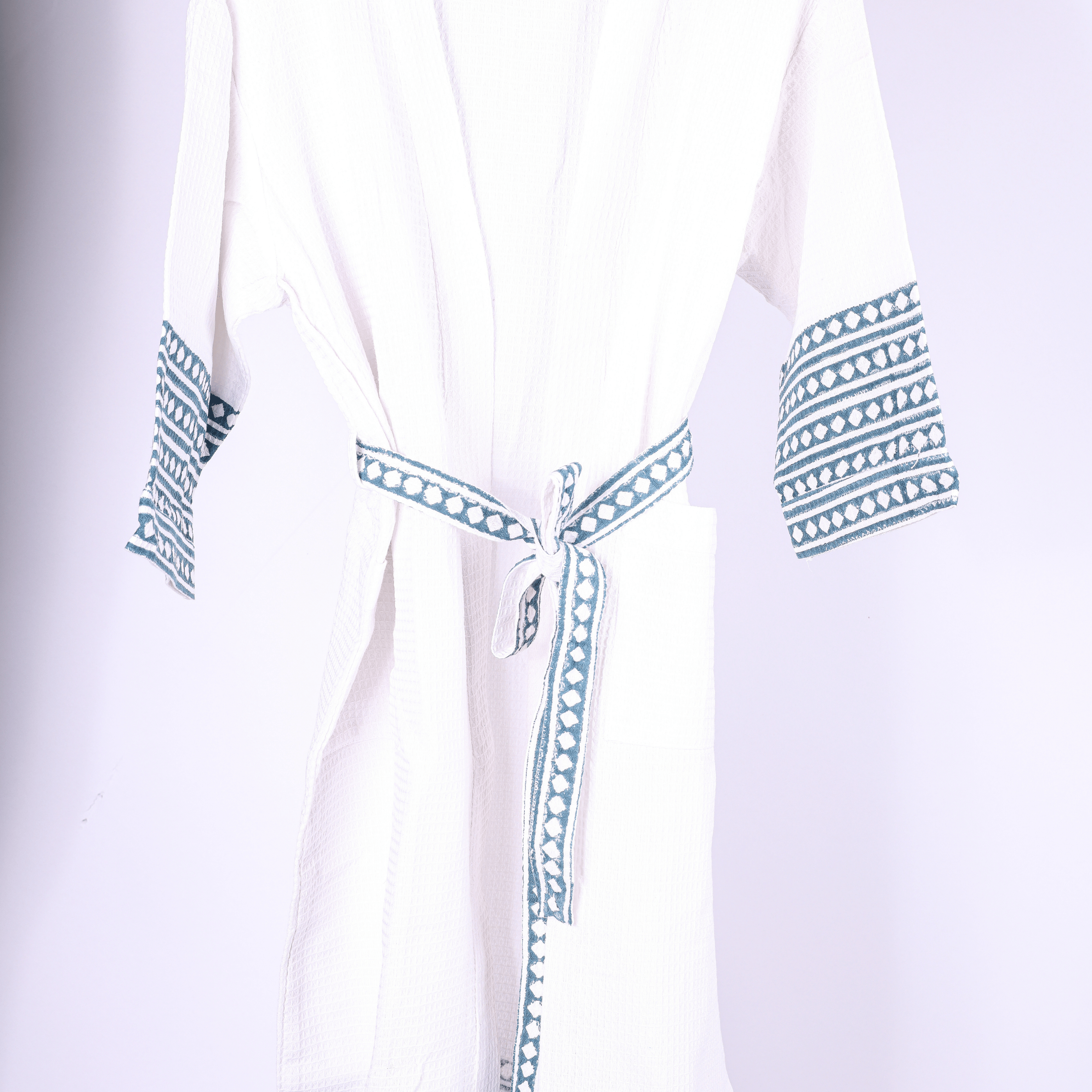 A white cotton bathrobe with blue geometric patterned trim on the cuffs, hem, and tie belt, hanging on a wooden hanger against a light background.