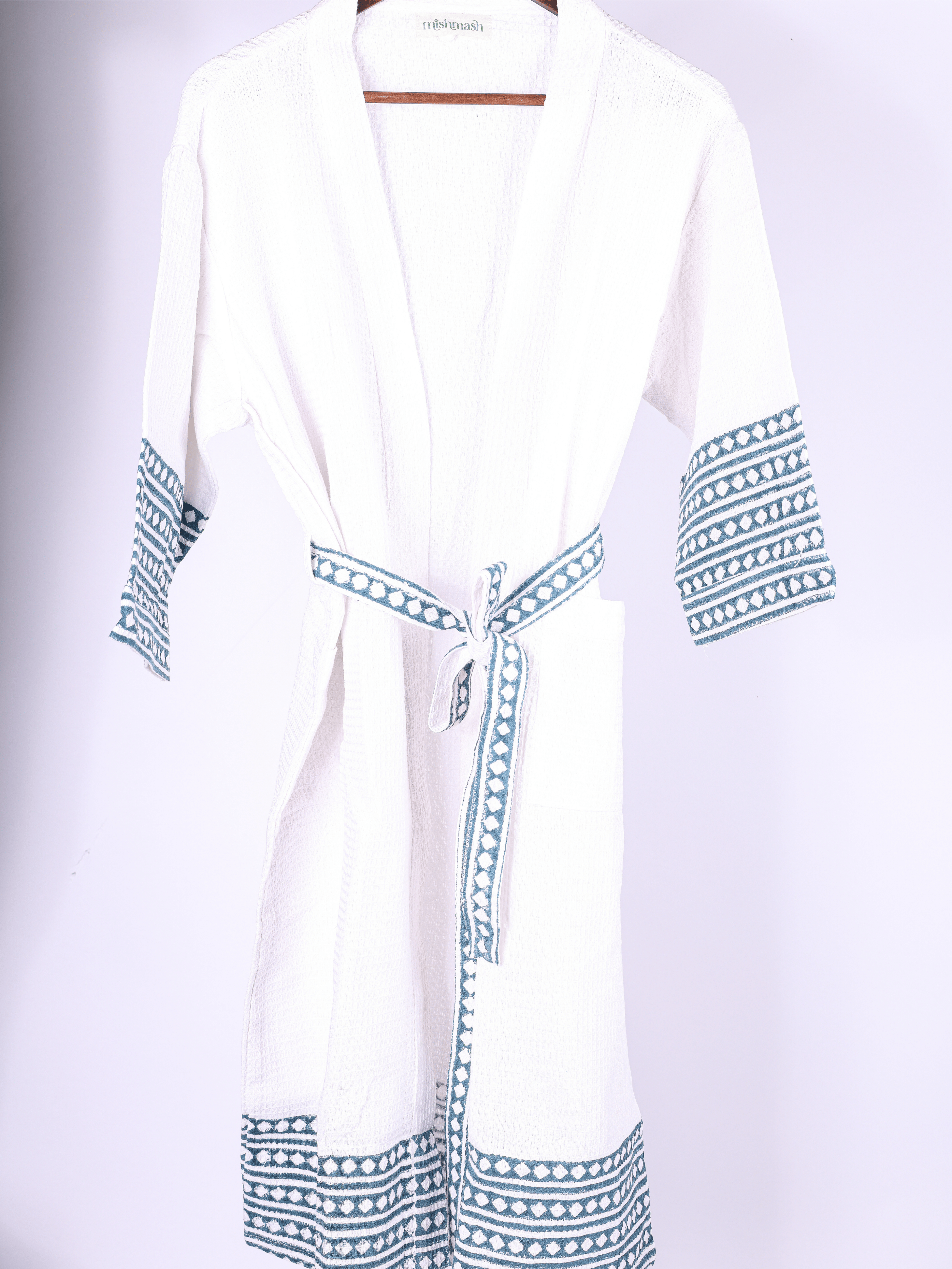 A white cotton bathrobe with blue geometric patterned trim on the cuffs, hem, and tie belt, hanging on a wooden hanger against a light background.